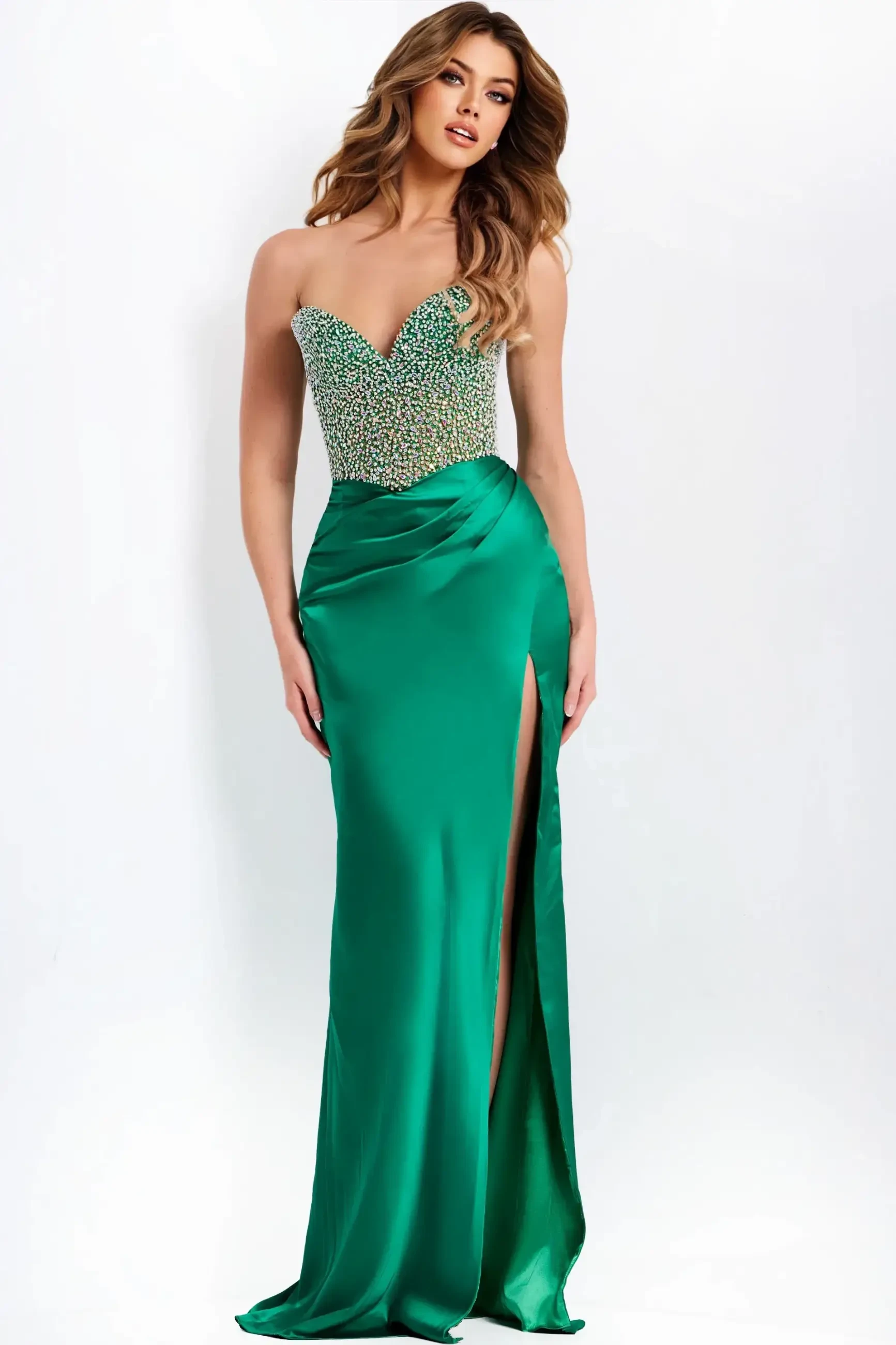 A woman in an elegant emerald green gown with a beaded, strapless bodice and a high slit stands confidently against a white background.