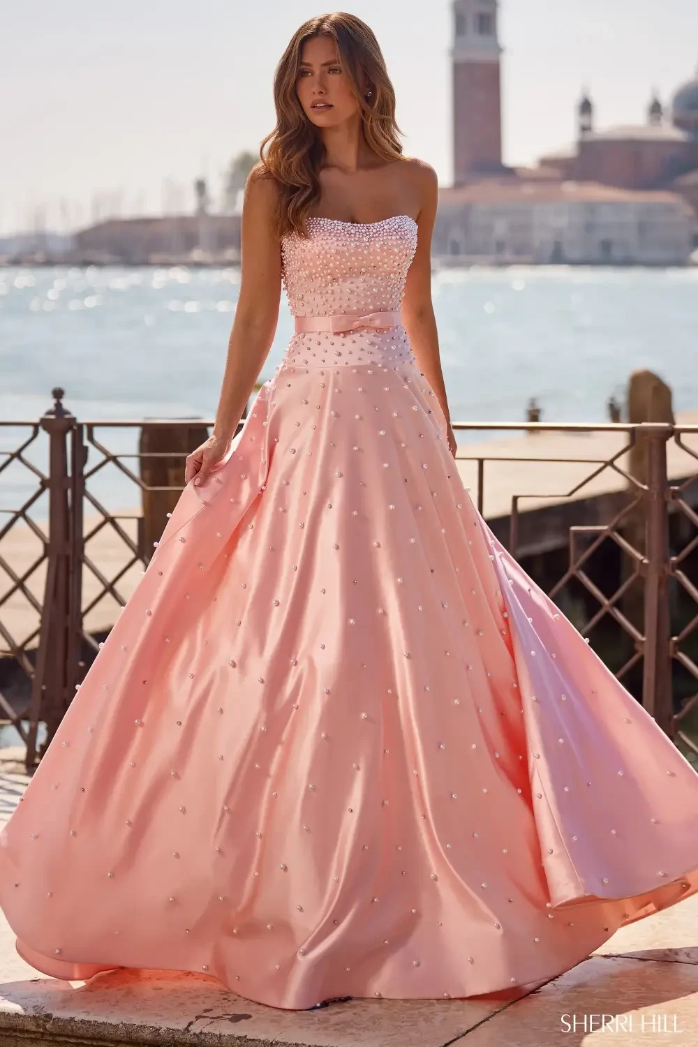 A woman in a strapless, pink gown adorned with pearls, standing by a waterfront with a scenic backdrop.