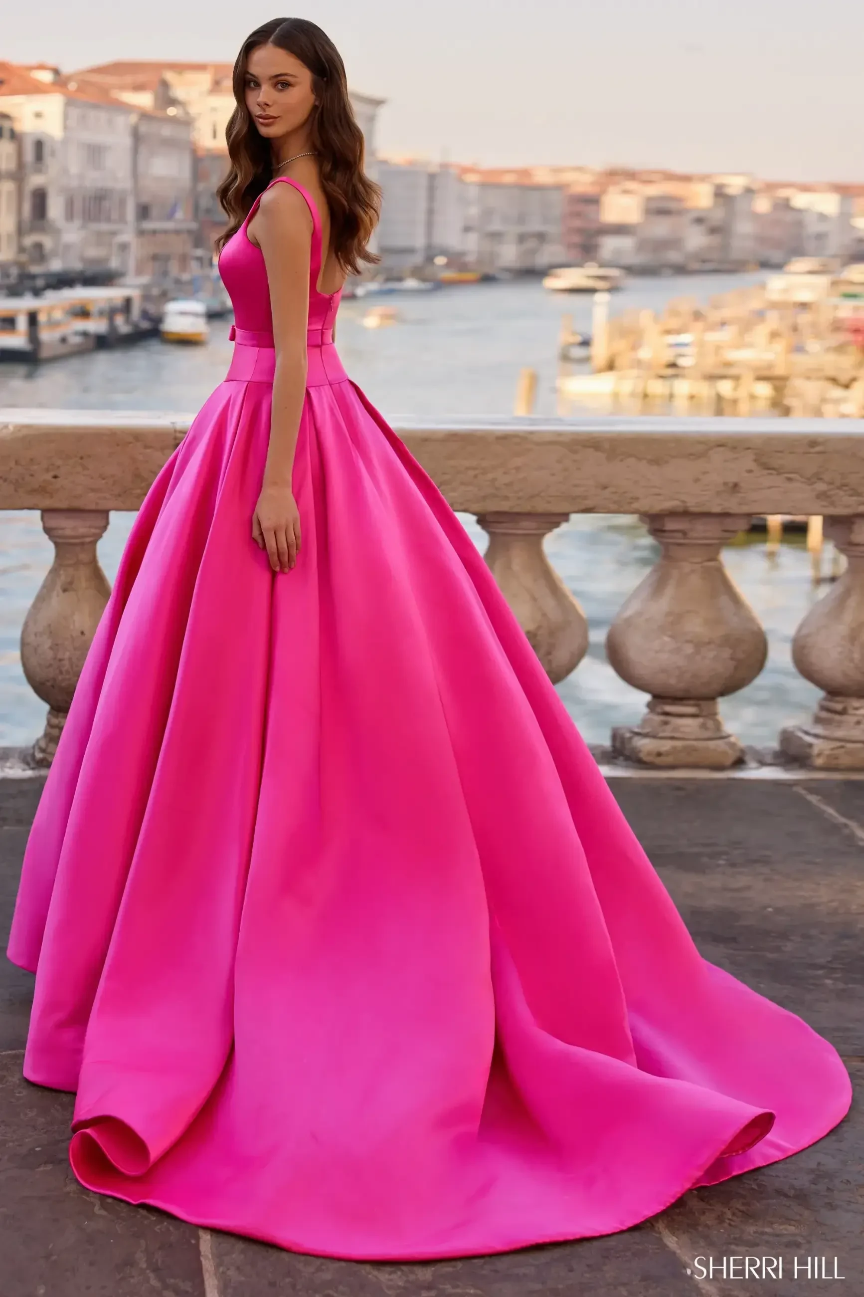 A woman in a vibrant pink ball gown stands by a stone railing overlooking a picturesque, sunlit canal. The scene conveys elegance and serenity.